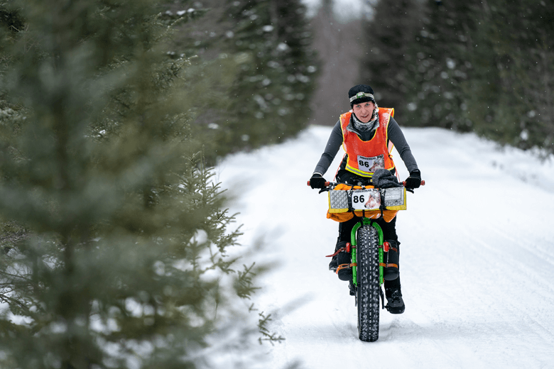 A front view of a winter cyclist riding a green fat bike on a snow covered trail between pine trees