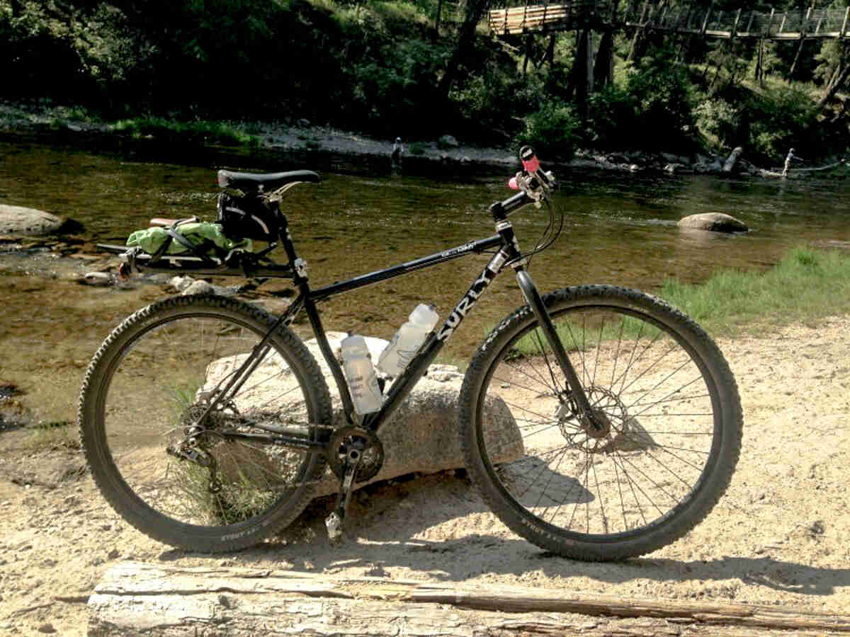 Right side view of a black Surly bike, parked against a rock on a riverbank, with a river and trees in the background