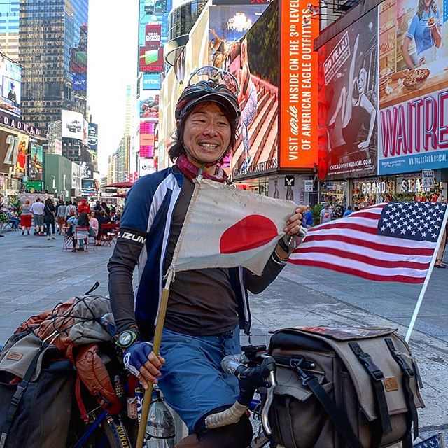 Cyclist standing with bike loaded with gear holds a Japan flag while on a busy city street with buildings all around