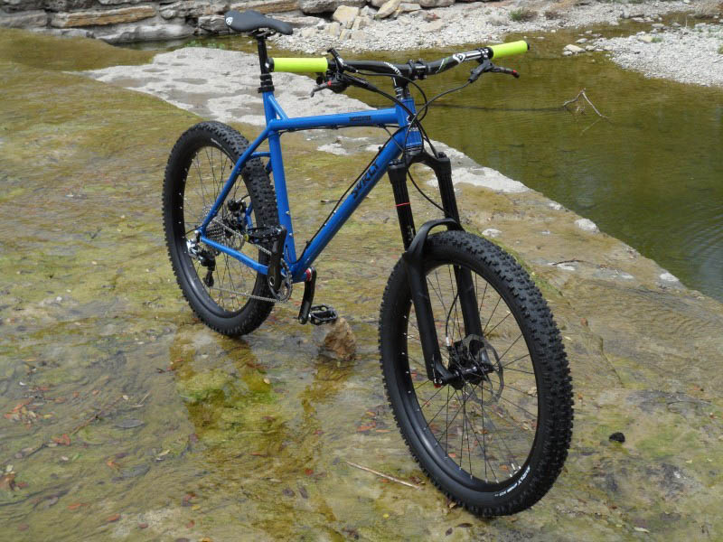 Front right side view of a blue Surly Instigator bike, parked on a rock ledge with a layer of water, and a stream below in the background