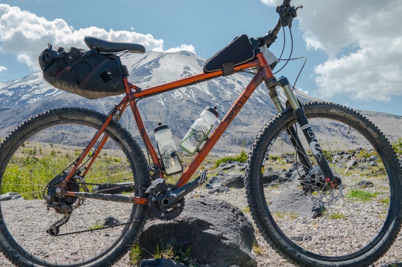 Right side view of an copper color Surly bike, parked on a rocky field, with a snow capped mountain in the background