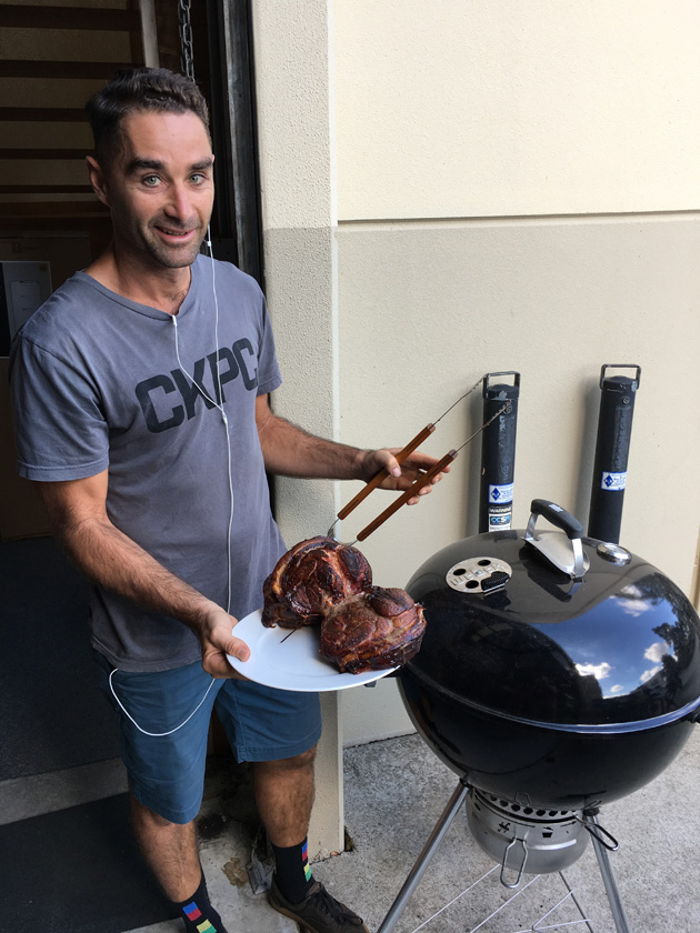 Person wear a gray t-shirt , stands next to a Weber grill holding a plate of meat 