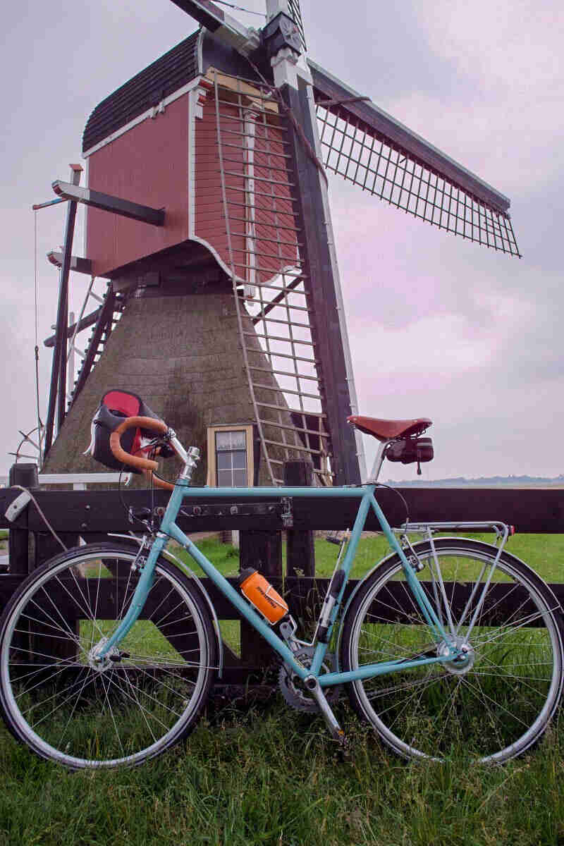 Left side view of a mint Surly bike, parked in a grass field, in front of a wooden fence, with a windmill in background