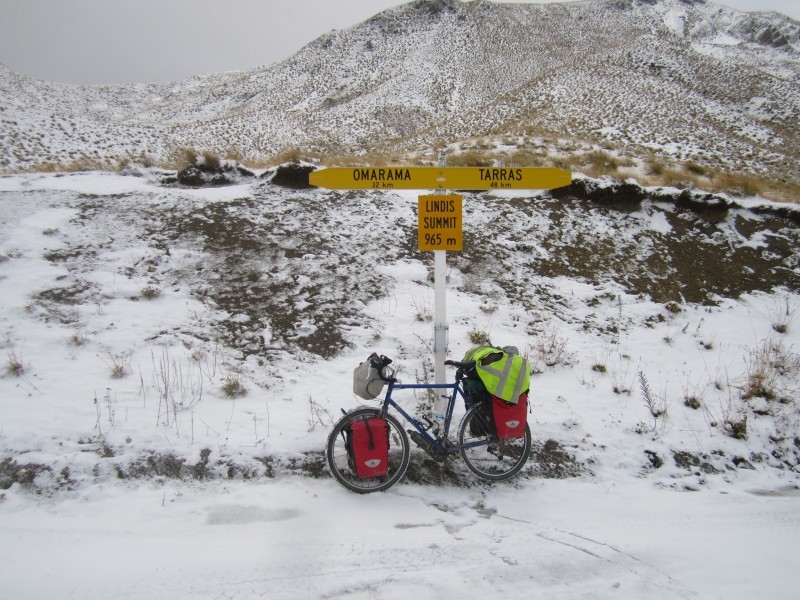 Left side view of a blue Surly bike with gear, parked in snow, against a directional sign at the base of a mountain