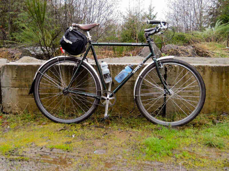 Right side view of Surly Cross Check bike with seat pack, parked in front of a short concrete wall with the woods behind