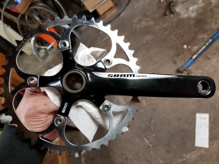 Downward view of a hand holding a right SRAM APEX bike crank with gear from below over a concrete floor