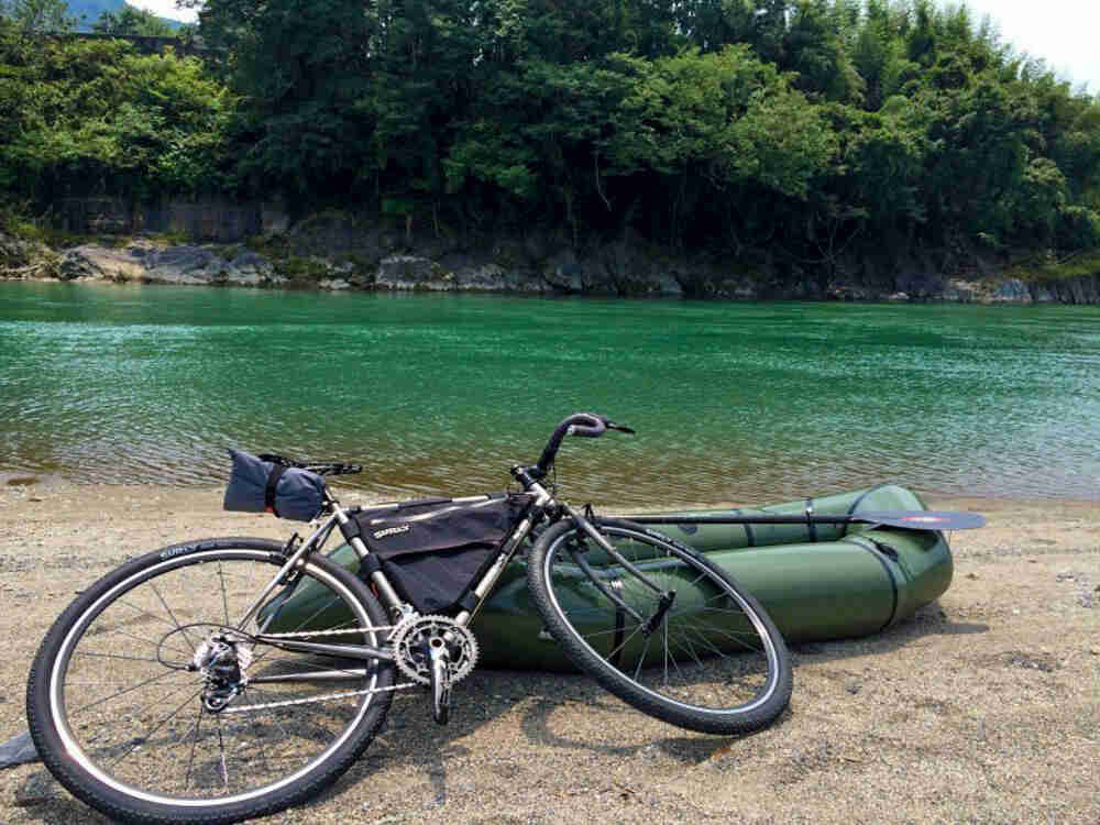 Right side view of a silver Surly Cross Check bike, lying on an inflated raft, on the shore of a turquoise lake