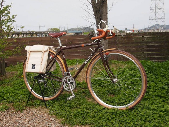 Right side view of a brown Surly Cross Check bike, parked on clover with a tree and wood fence in the background