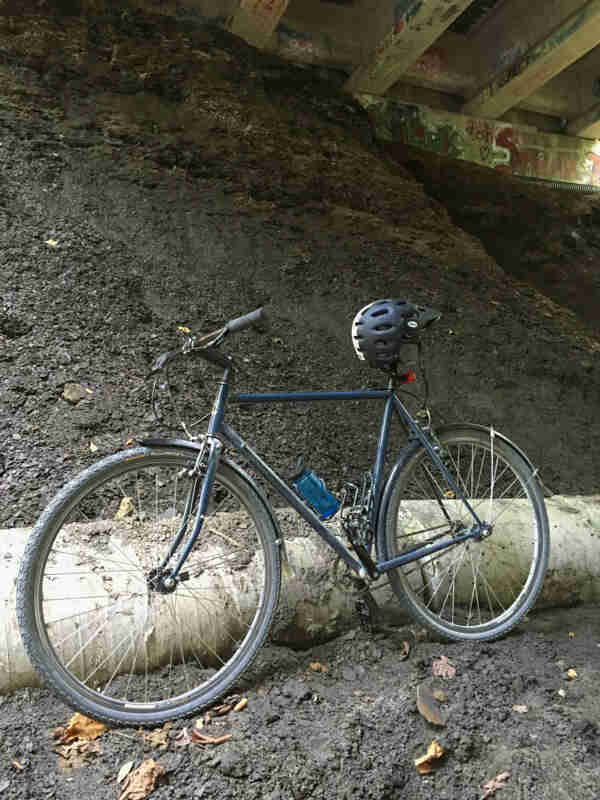 Left side view of a Surly Cross Check bike, with helmet on the seat, parked against a dirt hill, under a bridge overpass