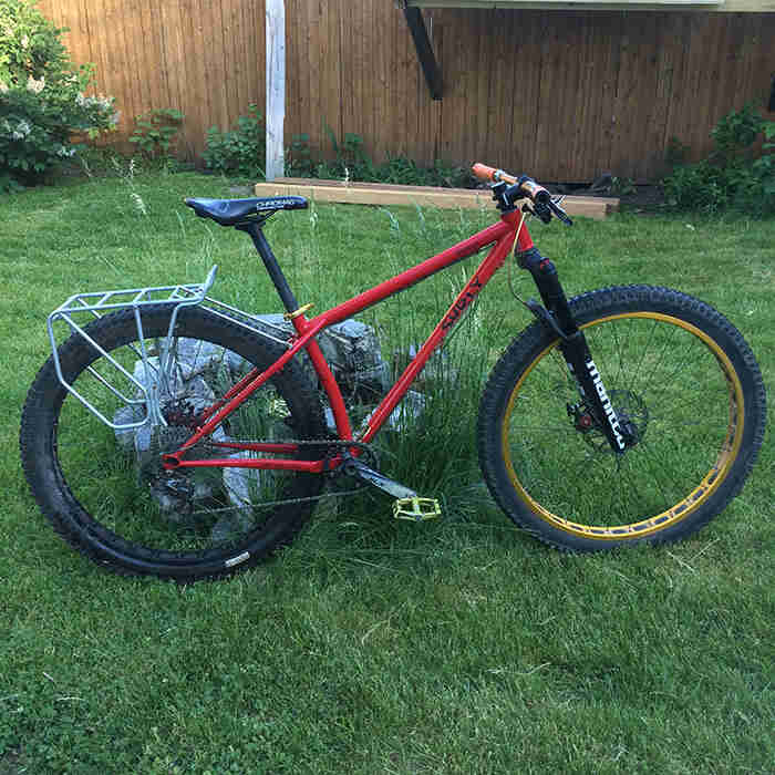 Right side view of a Surly Krampus bike, red, stand in a grassy yard against a rock, with a wood fence in the background