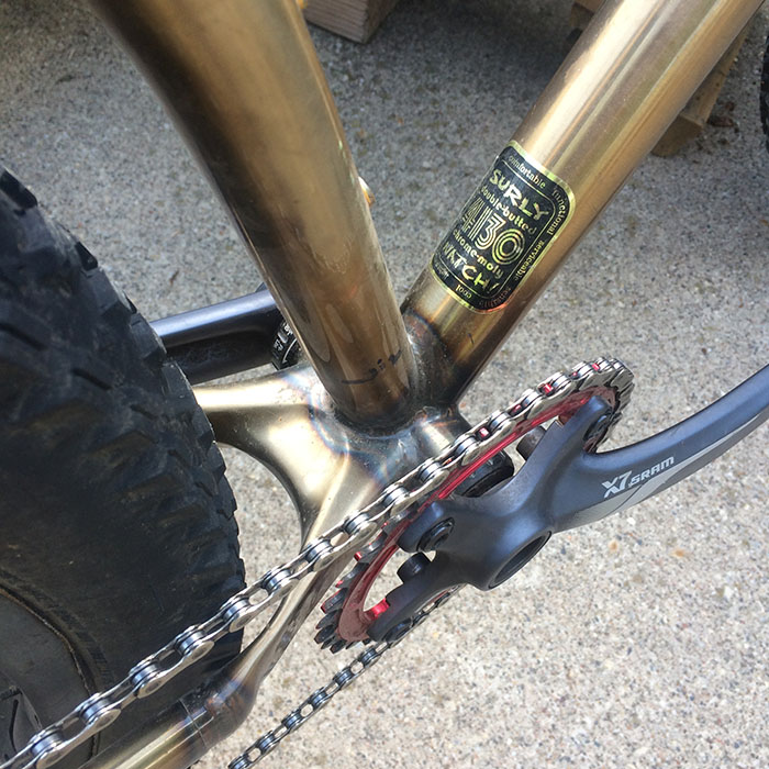 Downward close up view of a cranks and cast yoke on a Surly Krampus bike, gold, with pavement below