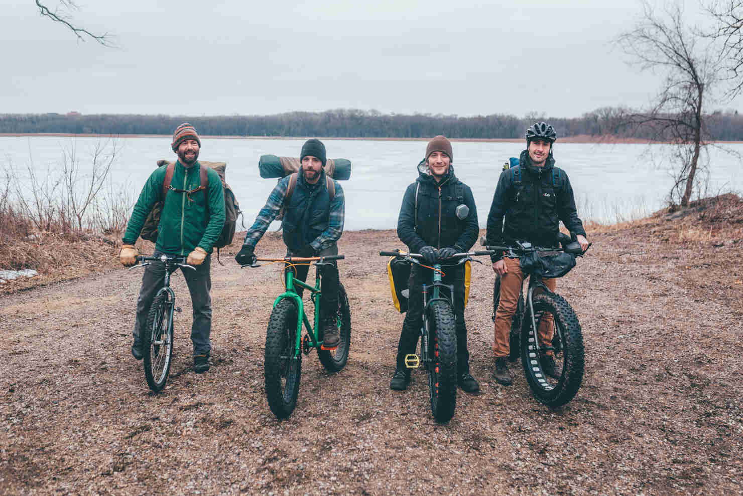 Front view of 4 cyclist facing straight ahead, stand on a gravel lot, with a frozen river in the background