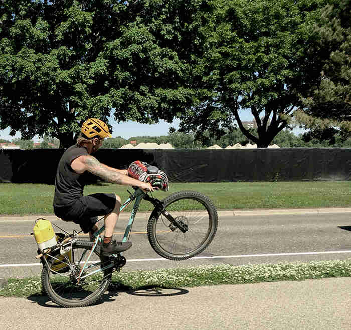 Right side view of a cyclist, on a Surly Wednesday bike, riding a wheelie down a sidewalk, with trees in the background