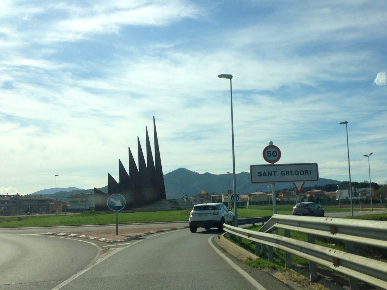 Rear view of a white SUV veering to the right, on a street, passing a Sant Gregori road sign, with mountains ahead