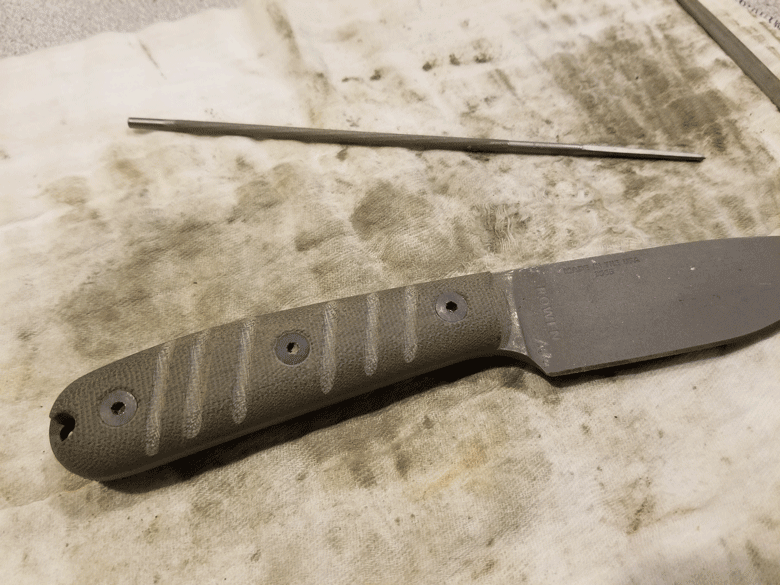 Gray fixed blade knife and round file laying horizontally on a dirty white towel