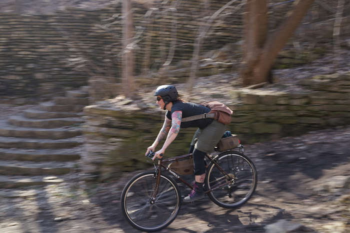 Left side of a cyclist riding a Surly Cross Check bike, down a hill, with a rock wall and trees in the background