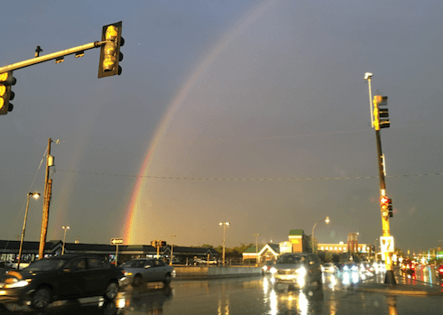 Part of a rainbow, arching over a wet street with traffic, with dark skies above