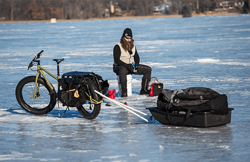 A person sitting on a bucket on a frozen lake, with a sled trailer attached to a Surly fat bike in front of them