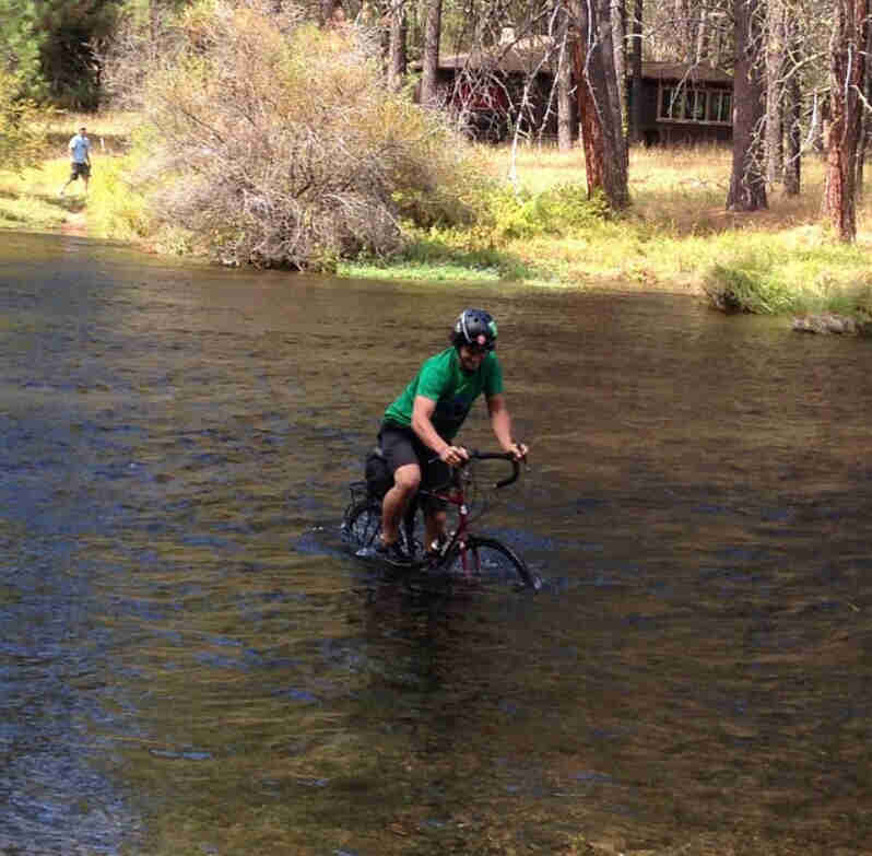 Front, right side view of a cyclist riding a red Surly bike through a river, with trees and a house in the background