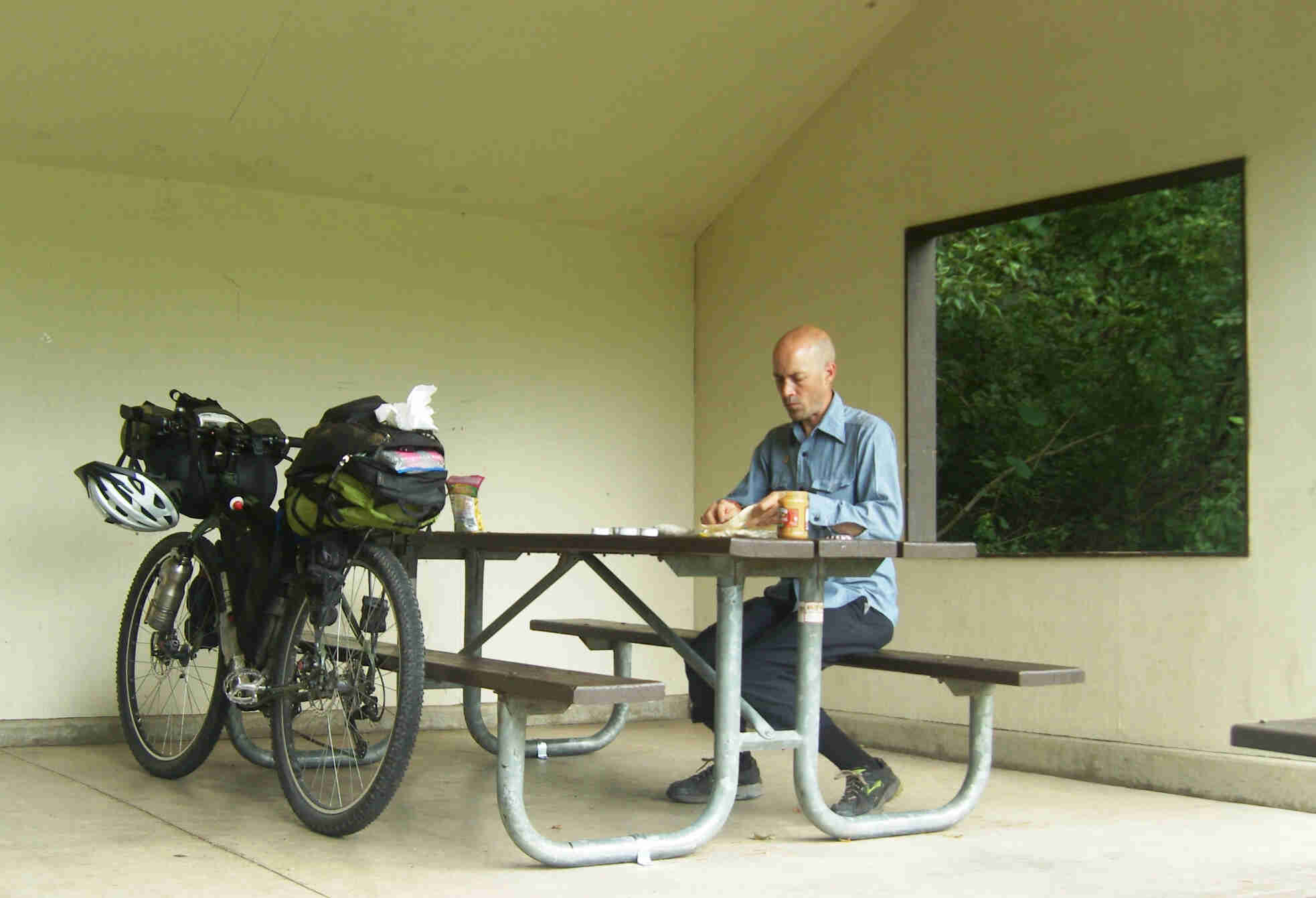 Person sitting at a picnic table, making a peanut butter sandwich, with a fully loaded bike parked on the other side