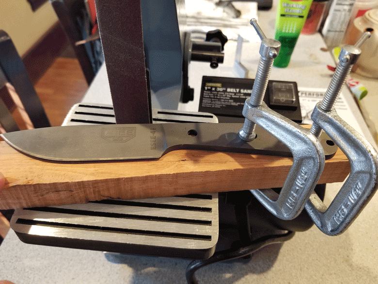 Downward view of a gray knife with no handle covers mounted to  board wtih two vises on a workbench
