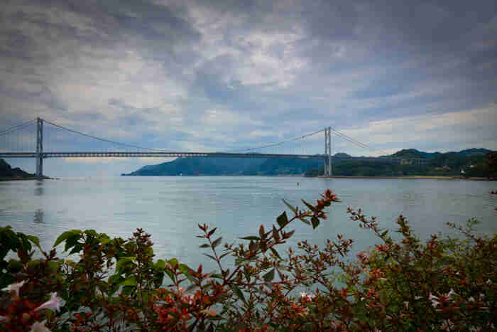 Side view of a suspension bridge, spanning over a bay, with tree covered hills behind it