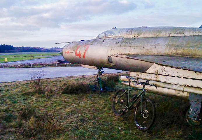Front, right side view of a green bike, parked on grass, and leaning on an old jet at an airfield