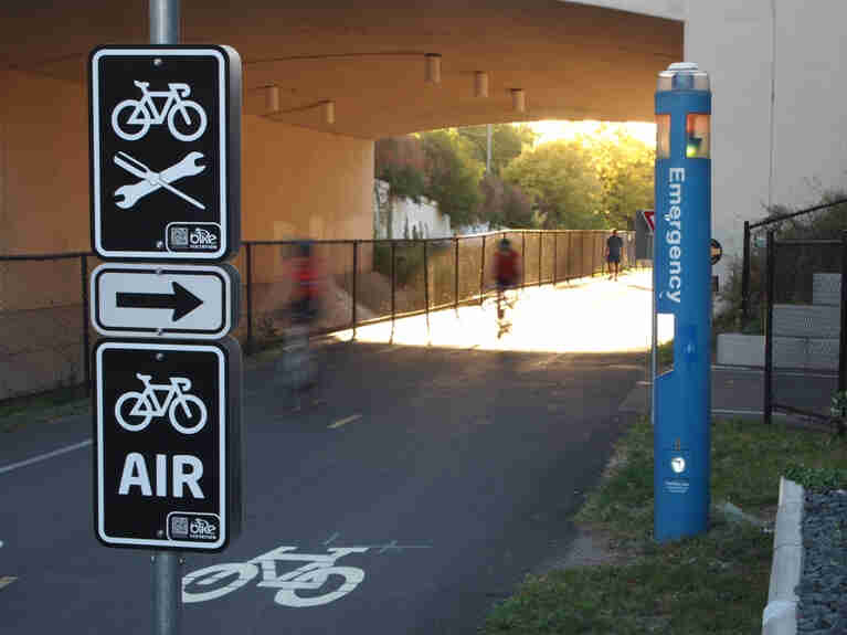 Road signs indicating bike repair and air available, with cyclists riding under a bridge tunnel