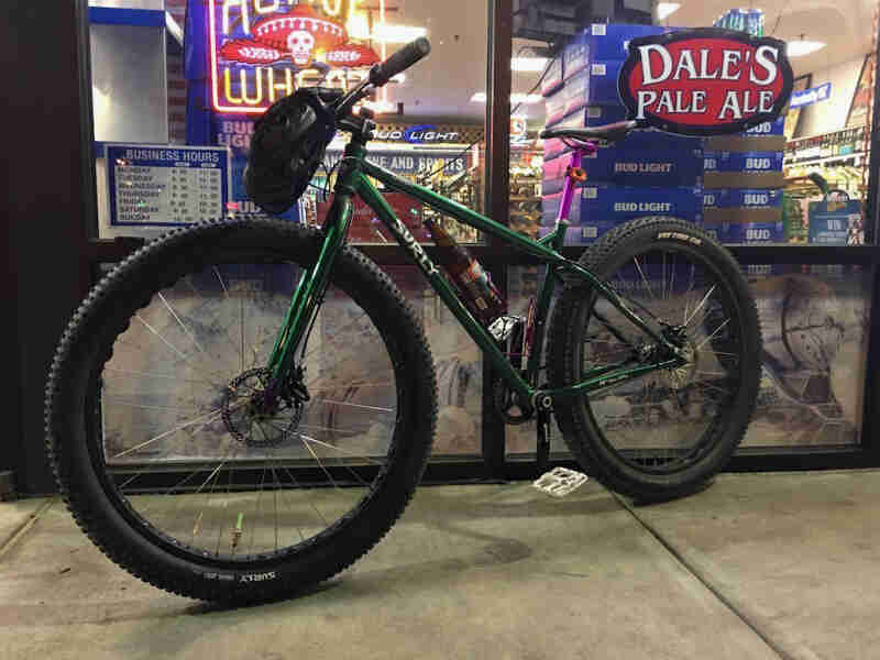 Left side view of a Surly bike, green, parked on sidewalk in front of a liquor store at night