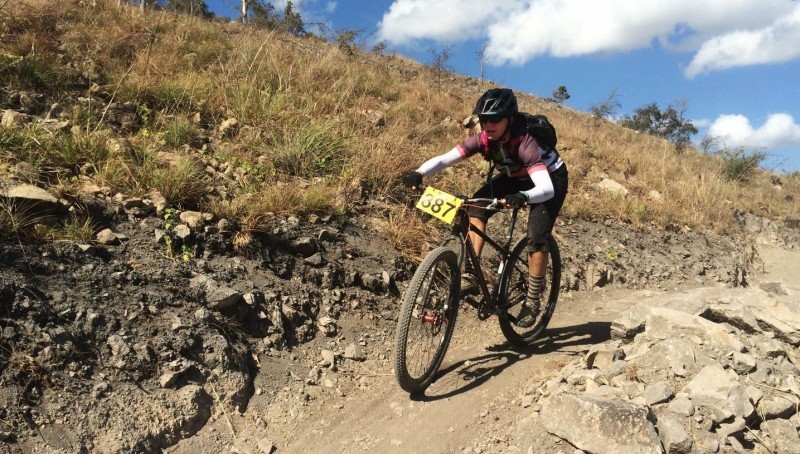 Front view of a cyclist, riding a Surly bike with a race tag in front, down a rocky trail on a grassy hill