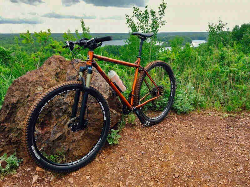 Left side angled view of a Surly bike, copper, parked on gravel in front of a rock, with weedy field in the background