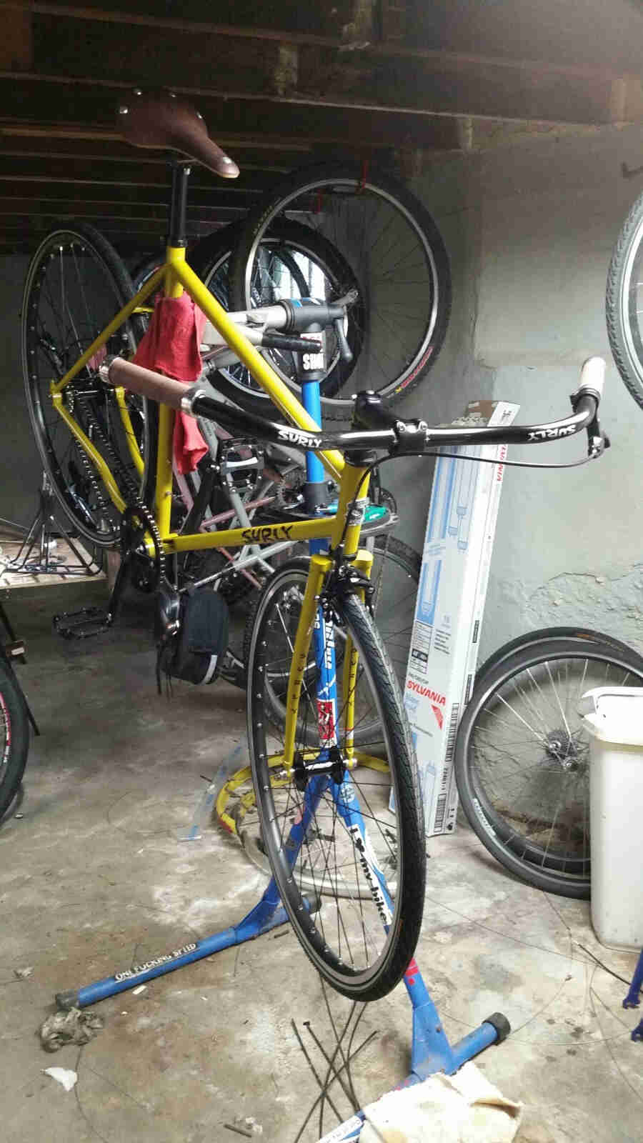 Front right view of a Surly Steamroller bike, yellow, on a bike stand, in a room with bike parts in the background