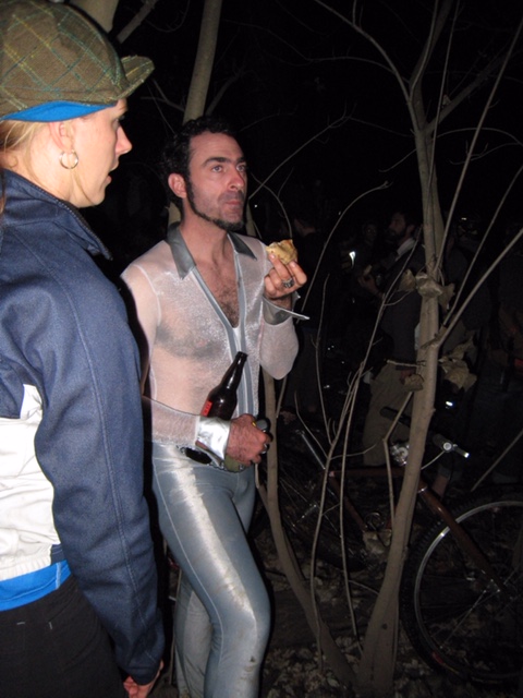 Front right side view of a person wearing a white bike race suit, in the woods at night