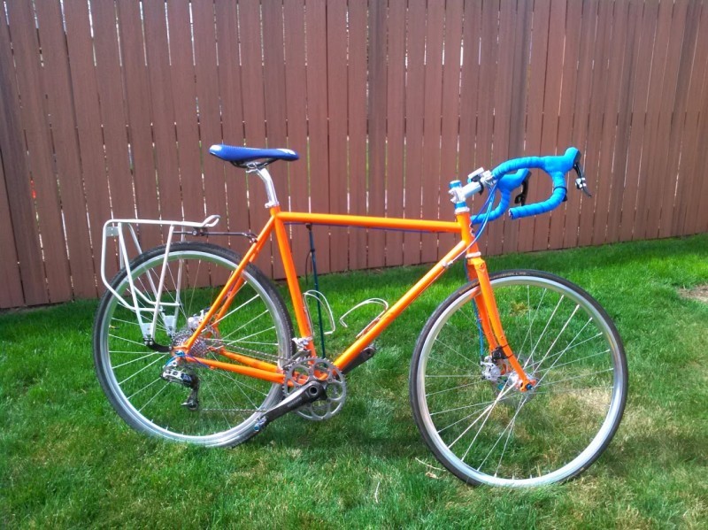 Right side view of an orange Surly Straggler bike, parked on grass in front of a wood fence wall