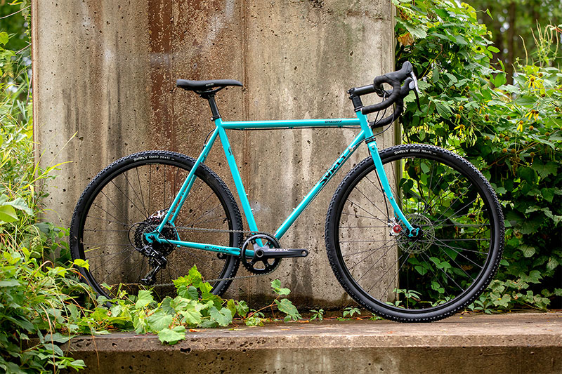 Blue color Surly Straggler complete bike side view leaning against large concrete road bridge support overgrown vines behind