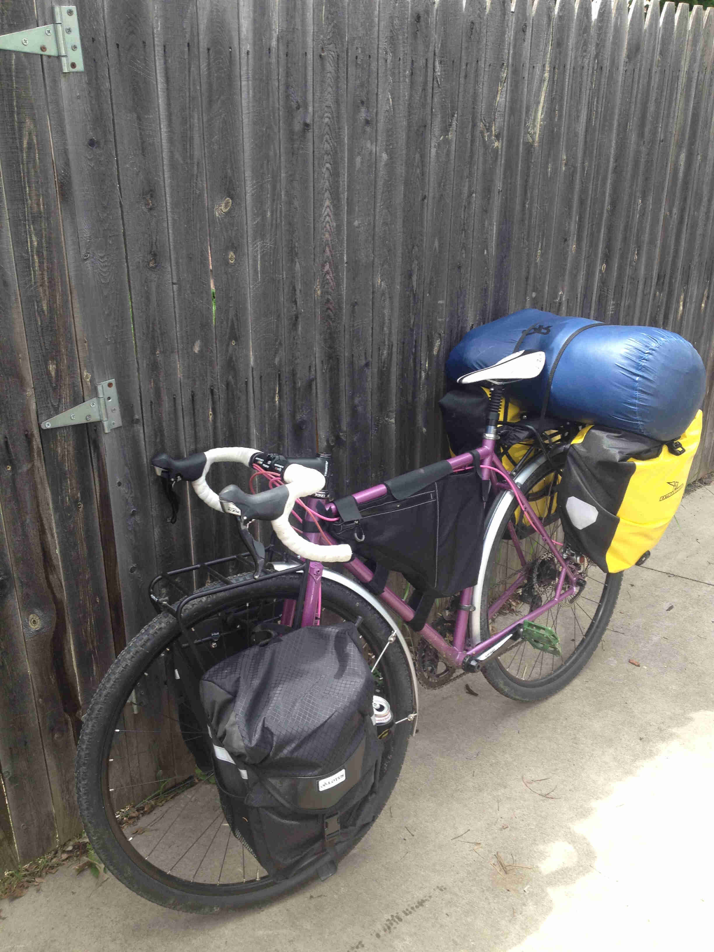 Left side view of a purple Surly Straggler bike, loaded with gear, leaning against a wood fence wall on a sidewalk