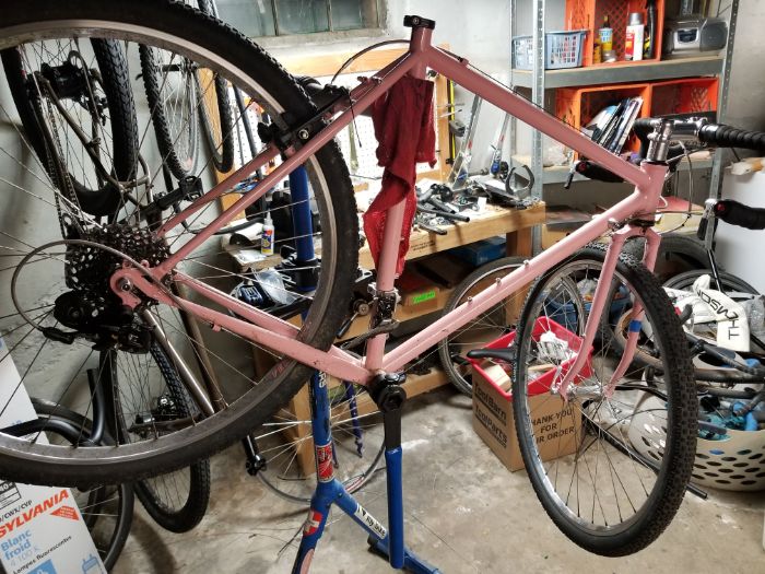 A bike with pink frame with no seat or post is mounted on a bike repair stand in a cluttered room with a concrete floor