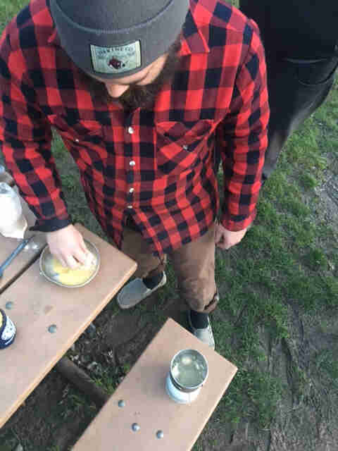 Downward view of a person, wearing a red flannel shirt,  mixing an egg in small bowl on top of a picnic table