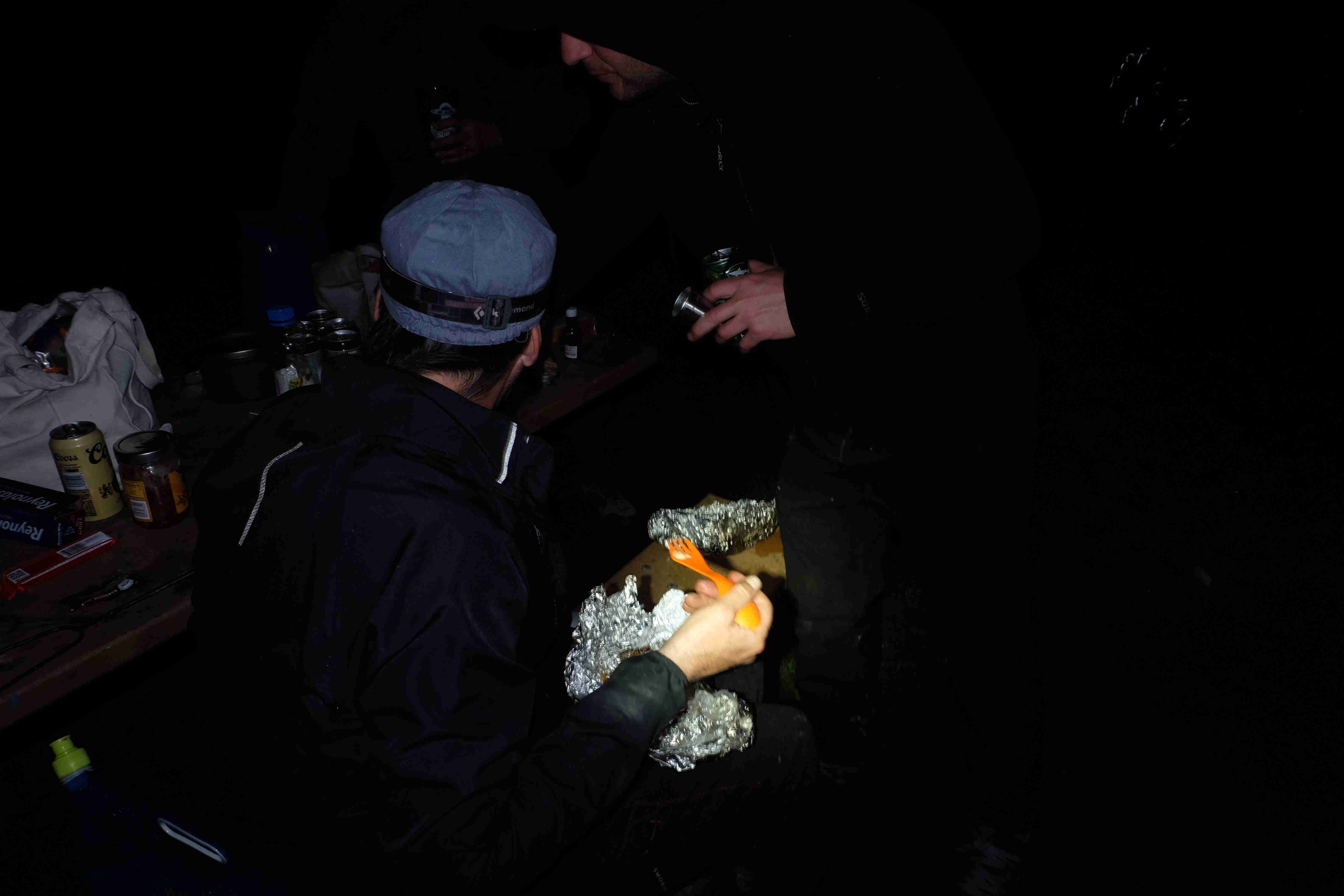 Two people at a picnic table, eating food on top of foil, in the dark at night