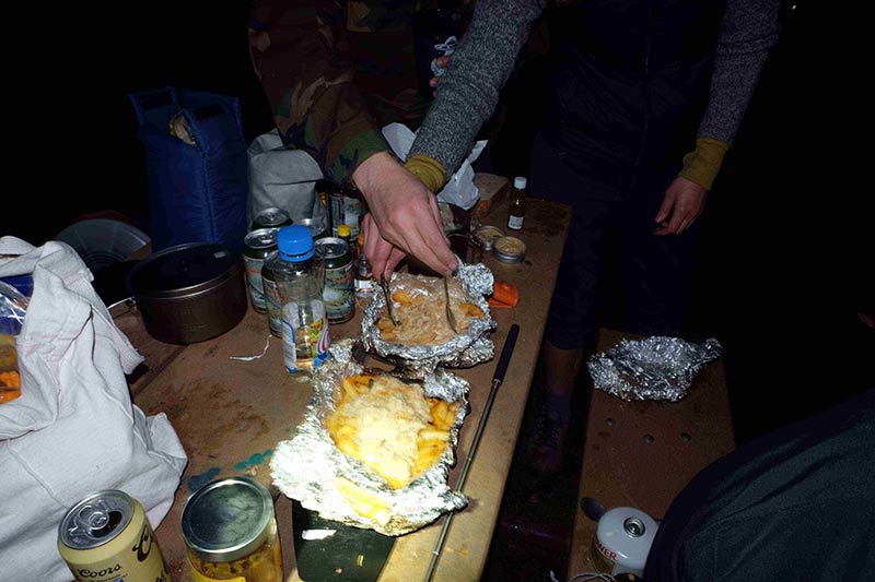 The hands of 2 people with forks, dig in to food sitting on foil, on top of a picnic table, in the dark at night