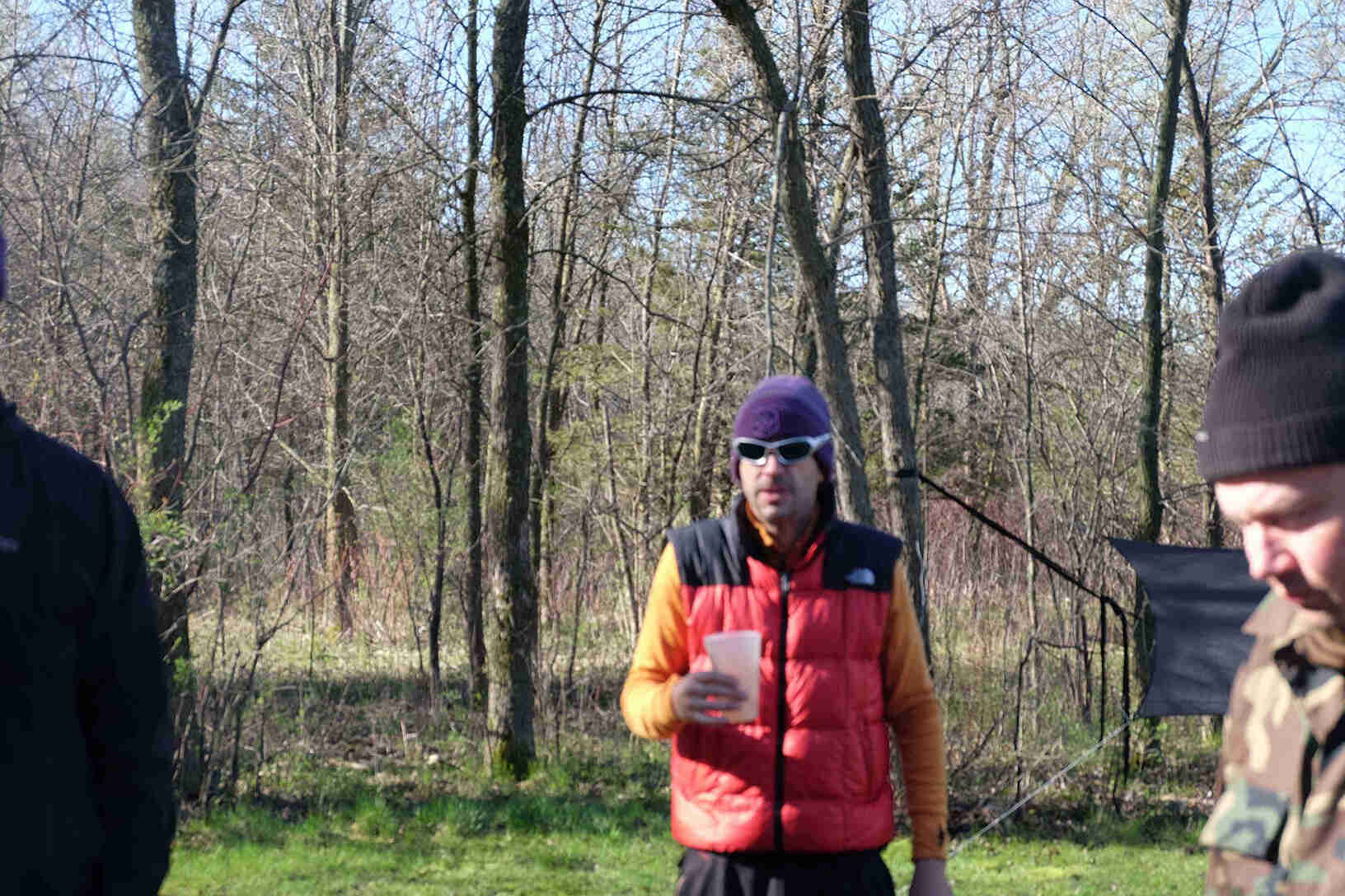 Front view of a person holding a plastic cup, wearing sunglasses, stocking cap and a vest, with trees in the background