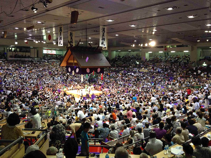 A view from the back of an arena, with spectators surrounding a sumo ring in the middle