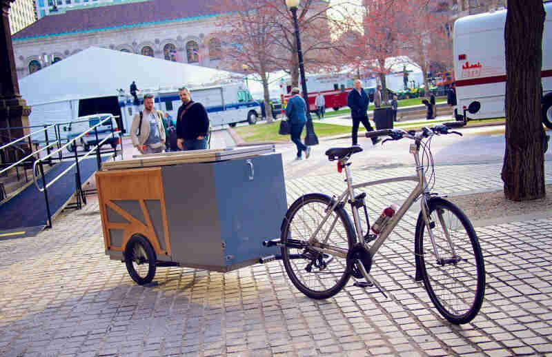 Front, angled view of a bike with a trailer, parked on a brick courtyard, with people standing behind