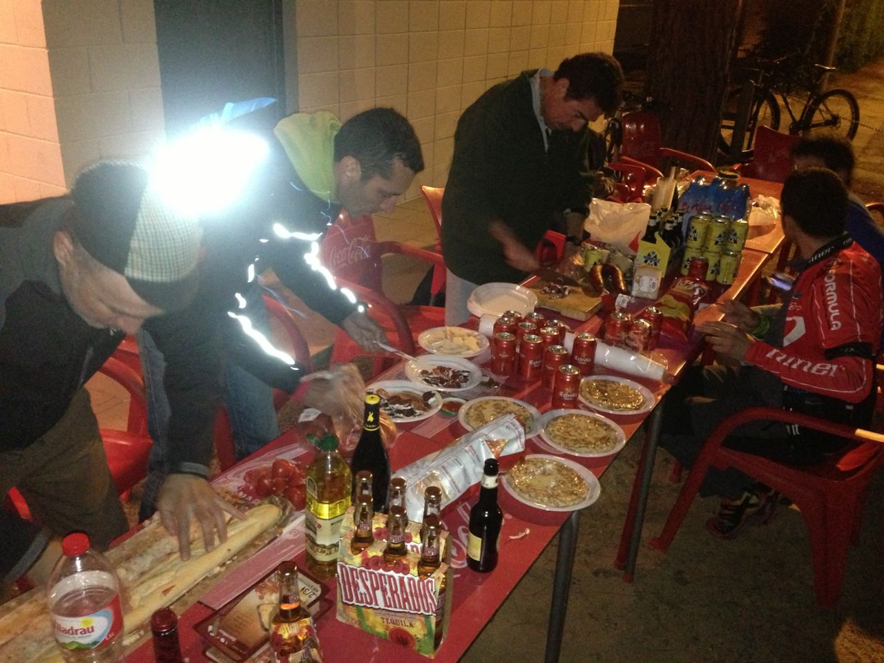 Side view of people, both sitting and standing, along a long table with food and beverages at night