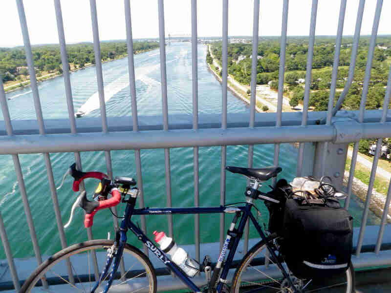 Left side view of a Surly bike, dark blue, parked along the rail on a bridge over a river, with trees on both sides