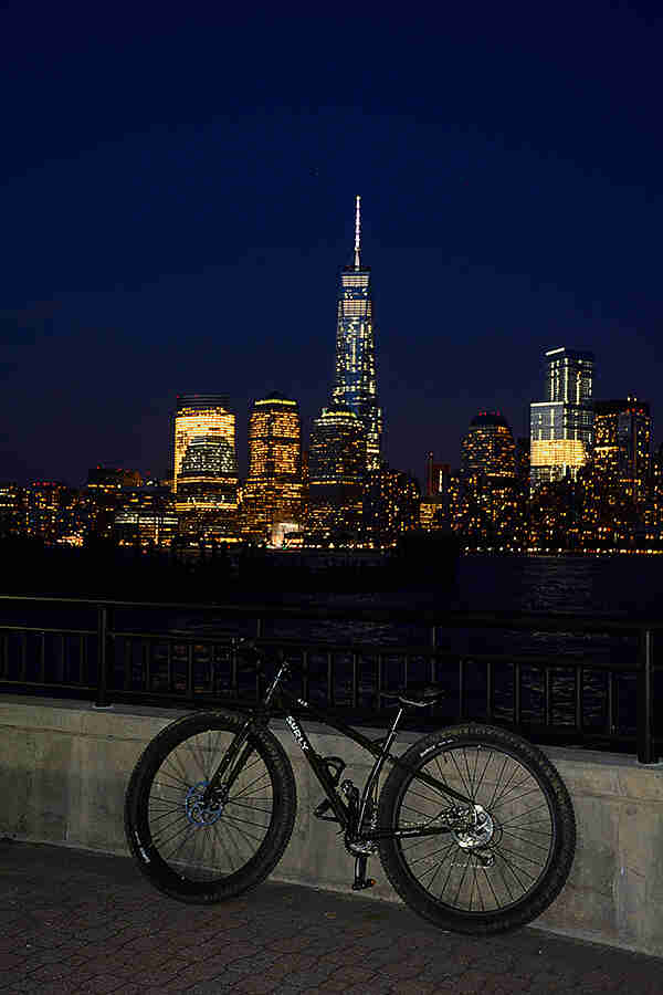 Left side view of a black Surly ECR bike, parked along a rail of cement boardwalk at night, with a city across the river