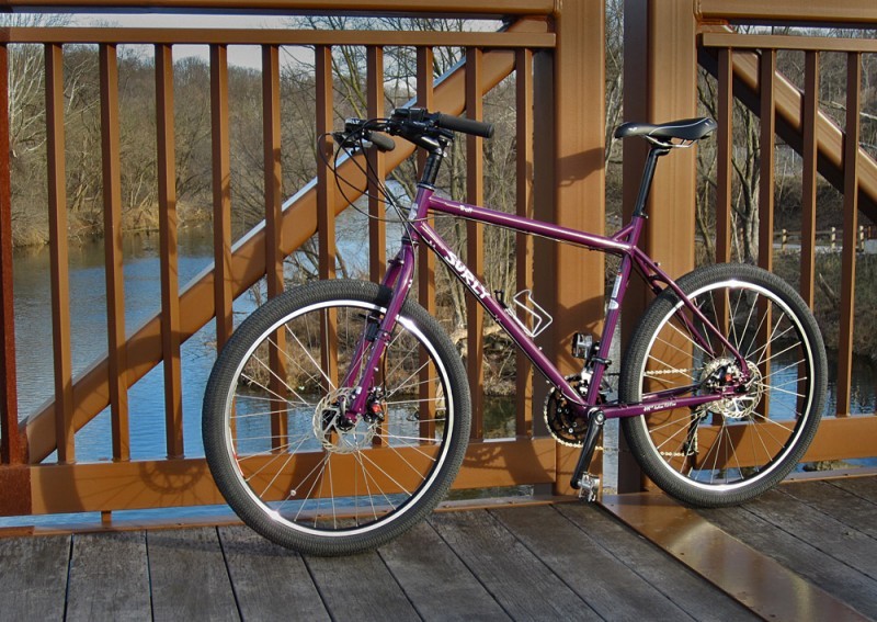Left side view of a purple Surly Troll bike, parked along the rail of a bridge that's over a river