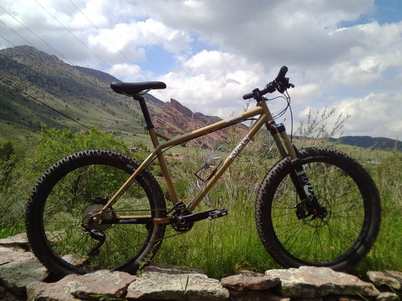 Right side view of a tan Surly Instigator bike, parked in grass on top of a rock wall, with mountains in the background