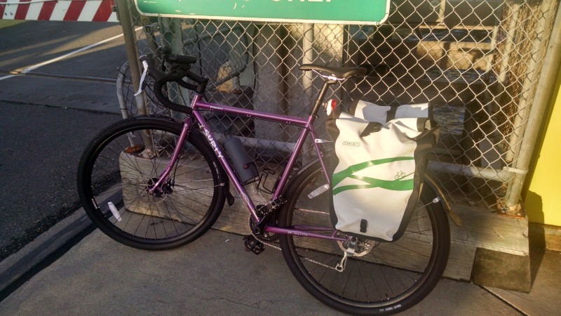 Left side view of a purple Surly Straggler bike, leaning against a chain link cage on a sidewalk, facing the street