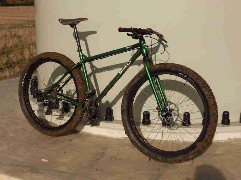 Right side view of a green Surly bike with muddy tires, parked on concrete and leaning against the base of a windmill